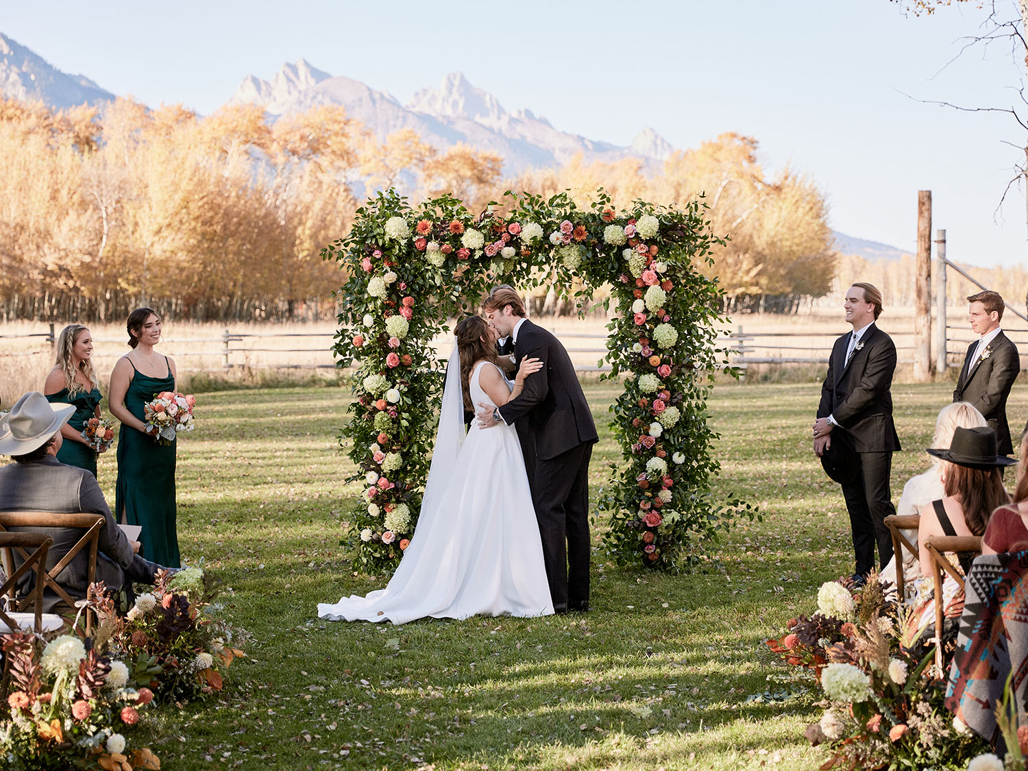 bride and groom kissing under flower arch with bridal party standing either side